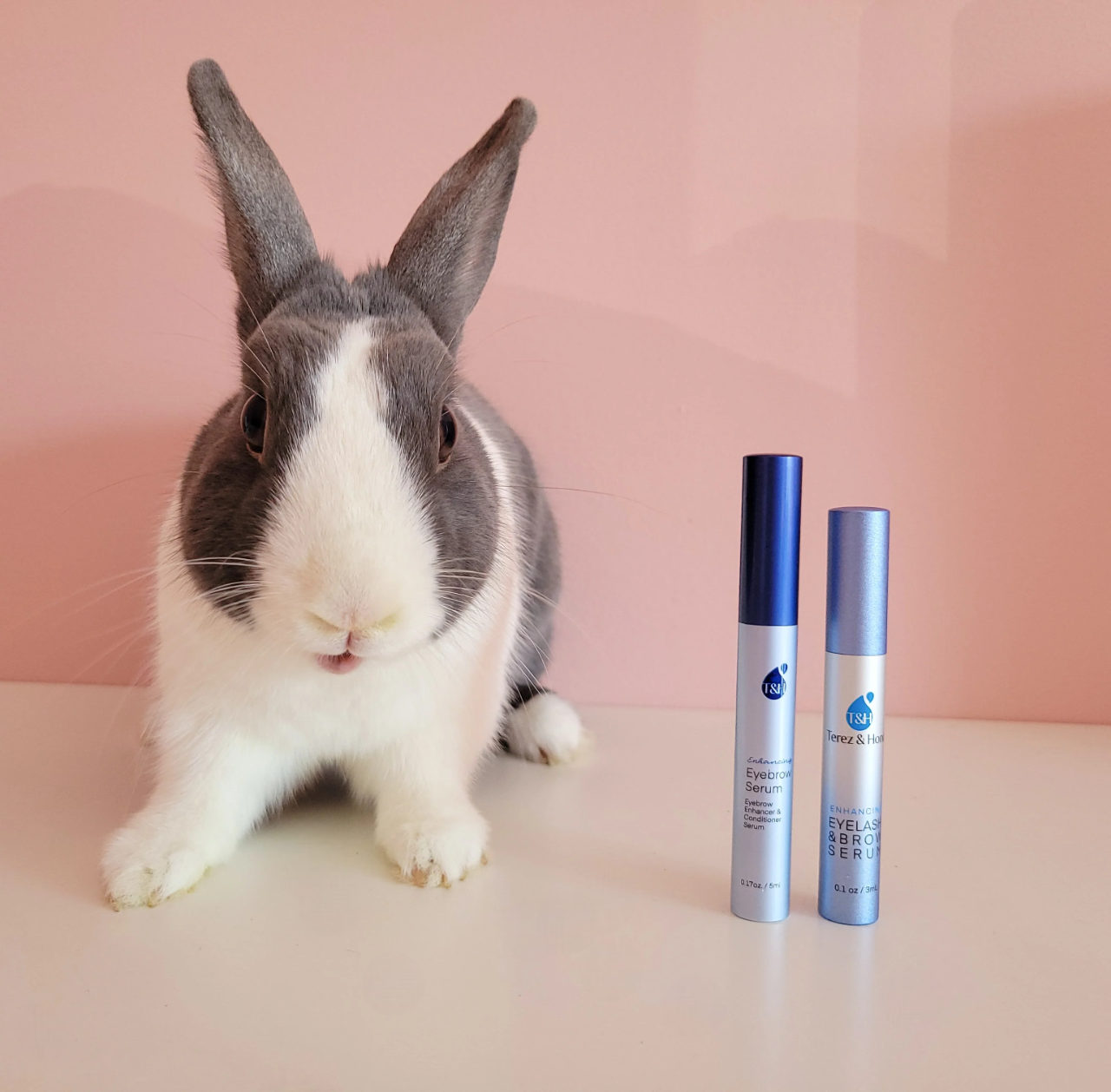 Adorable grey and white rabbit sitting beside two bottles of Terez & Honor Eyelash & Brow Serum on a pink background, highlighting the brand's cruelty-free commitment